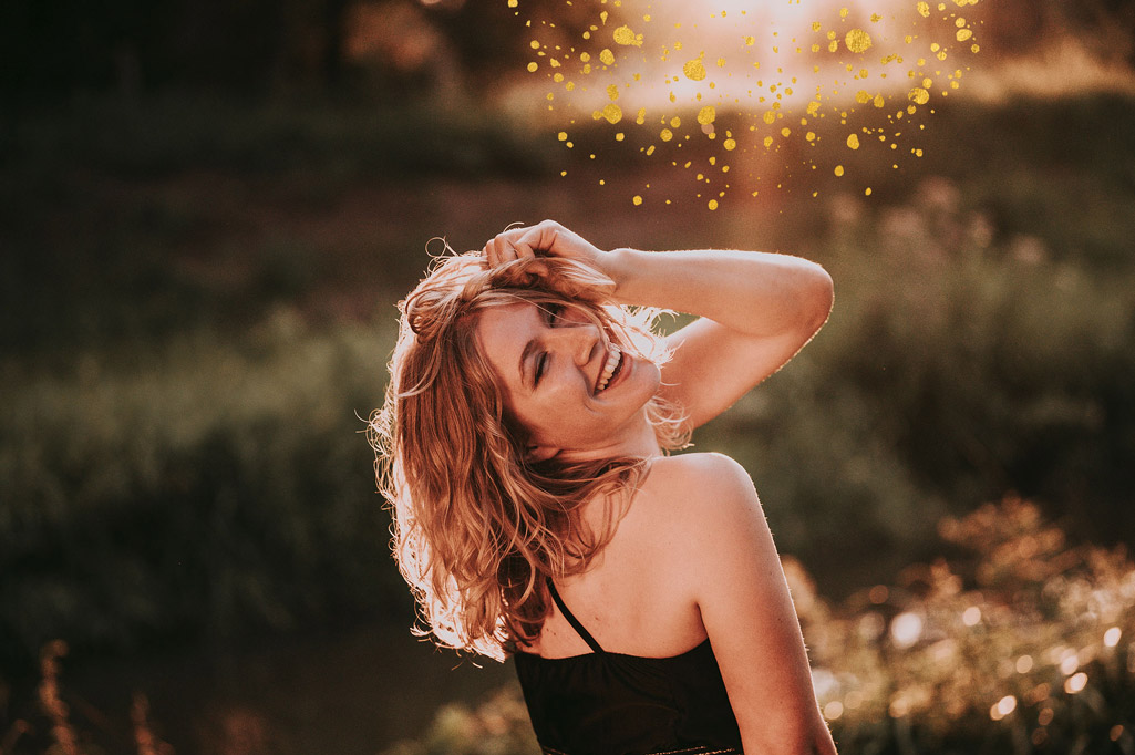 Johanna lachend in warmem Licht der Abendsonne auf einer Wiese. Der Hintergrund ist verschwommen. Dekorative goldene Punkte auf dem Bild.