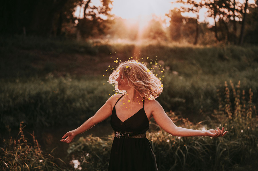 Johanna tanzt im schwarzen Kleid in der Abendsonne auf einer Wiese. Der Hintergrund ist verschwommen. Dekorative goldene Punkte schwirren um ihren Kopf.
