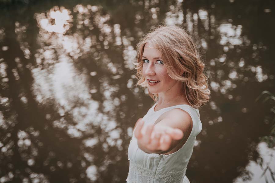 Johanna streckt dem Betrachter eine Hand entgegen und schaut direkt in die Augen: folge mir. Dahinter verschwommen dunkle Wasseroberfläche.